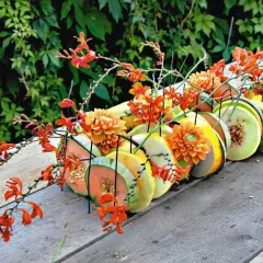 Letnia kompozycja z pomarańczowymi kwiatami i plastrami owoców, jak melon i ananas, na drewnianym stole.