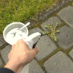 Ręka trzyma opryskiwacz, przygotowując się do zastosowania środka na chwasty. Obok stoi pojemnik z białym proszkiem i łyżeczką.