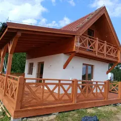 Nowy biały dom z drewnianym gankiem i balkonem, w otoczeniu zieleni.