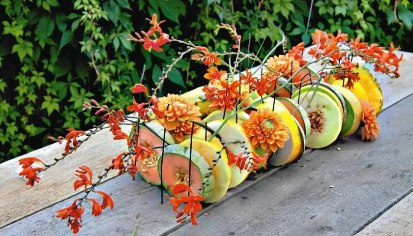 Letnia kompozycja z pomarańczowymi kwiatami i plastrami owoców, jak melon i ananas, na drewnianym stole.