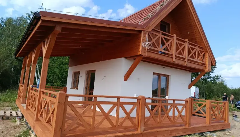 Nowy biały dom z drewnianym gankiem i balkonem, w otoczeniu zieleni.