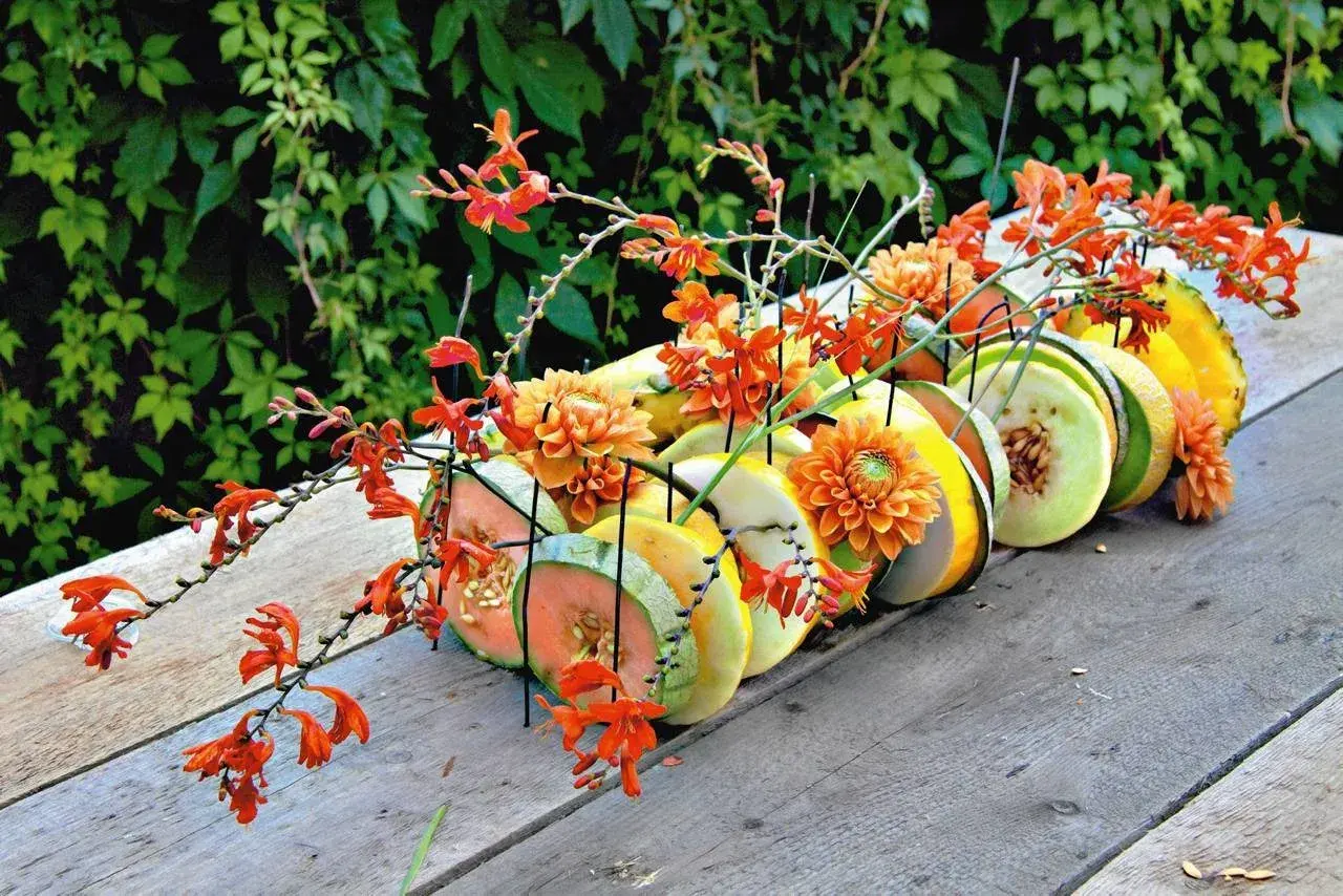 Letnia kompozycja z pomarańczowymi kwiatami i plastrami owoców, jak melon i ananas, na drewnianym stole.